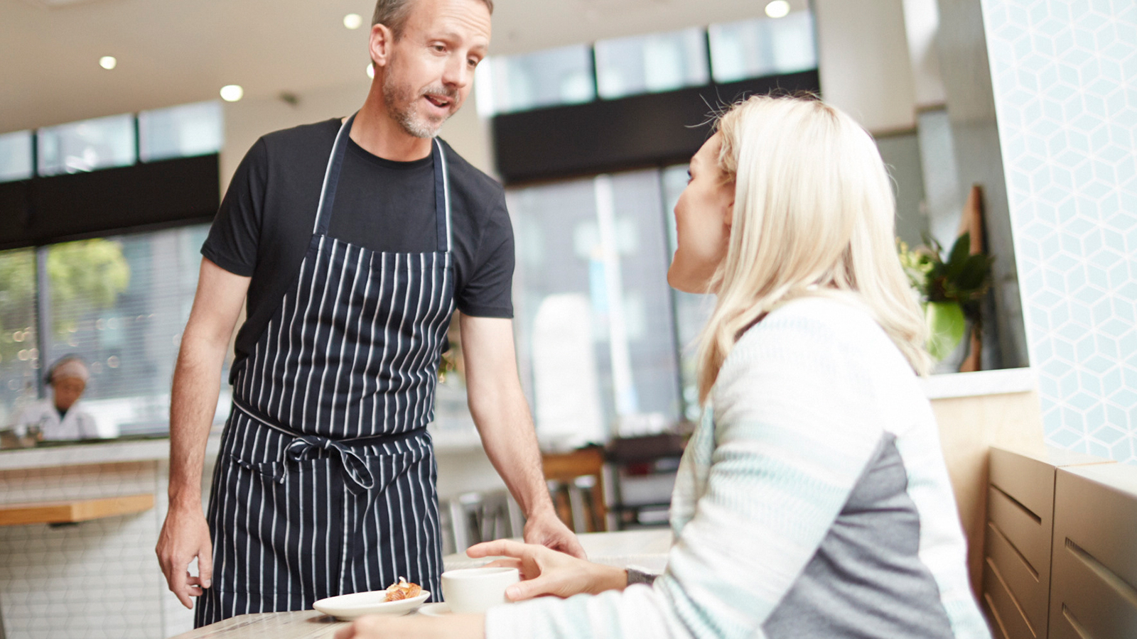 Mesonero hablando con cliente A man talking to a woman seated at a table in a café.