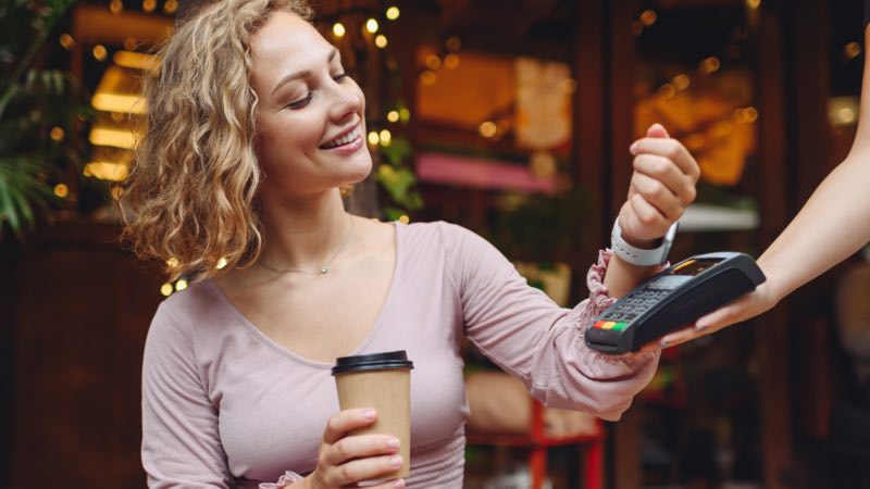 Imagem exibe mulher sorridente realizando pagamento por aproximação com smartwatch.